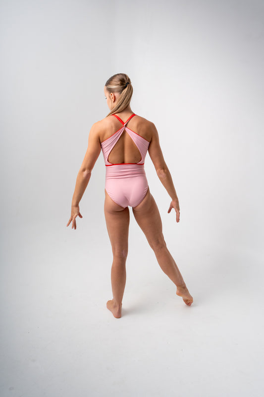 A dancer wearing a pink leotard with a cut-out back and red straps, viewed from the back, standing in a graceful pose.