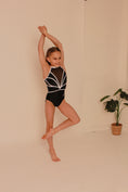 Carica l'immagine nel visualizzatore della galleria, A young girl in a black leotard with white accents, striking a dynamic pose with her arms raised and one leg lifted
